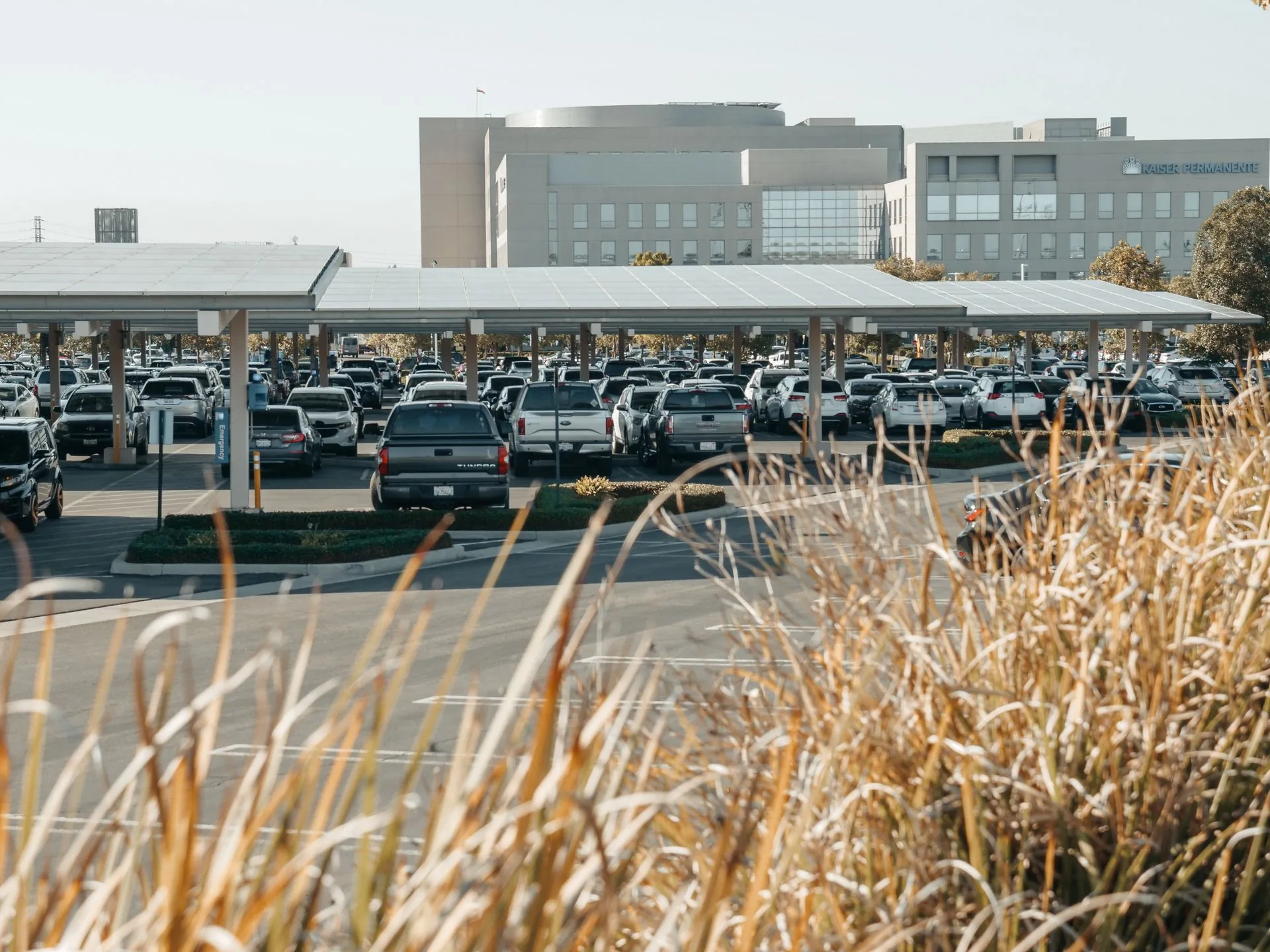 Overhead aerial view of long parallel rows of commercial solar carport canopies covering a parking lot, casting diagonal shadows across the asphalt, with cars parked along the edges and a roadway running alongside.